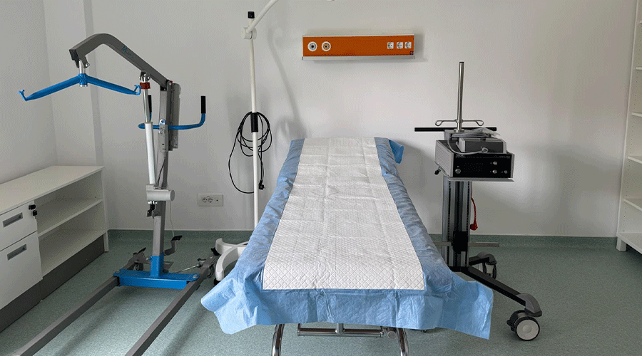 Operating room with an empty patient table covered in a blue sterile drape, a robotic arm on the left, and a mobile instrument cart on the right.
