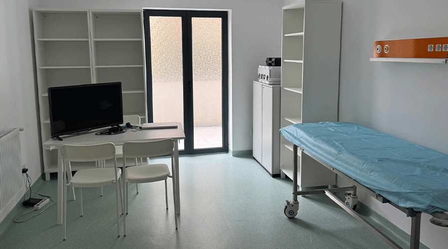 Small clinical examination room with a blue-covered exam table on wheels, a desk with a monitor and chairs, and tall white shelves near a glass door.