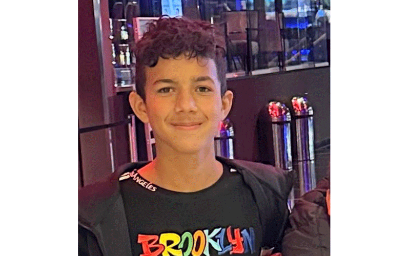Smiling boy with short dark hair in a black Brooklyn shirt, standing indoors near arcade machines or entertainment venue.