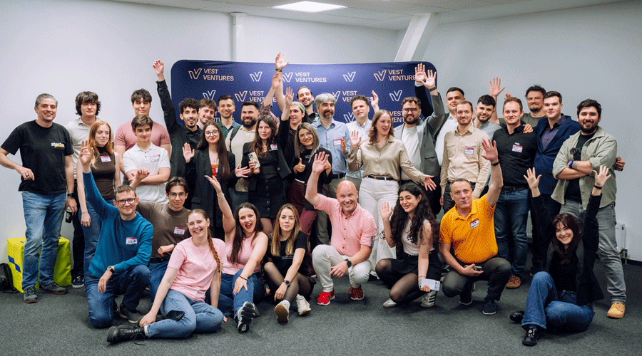 Large group of diverse people posing and waving in a conference room, with a Vest Ventures banner in the background.