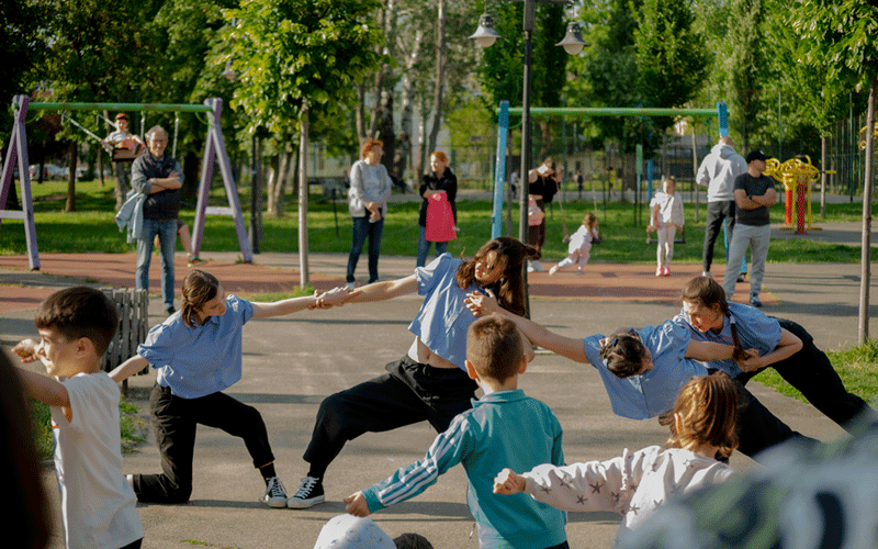 Group of children in blue shirts holding hands and stretching in a sunny park while adults watch nearby in the background.