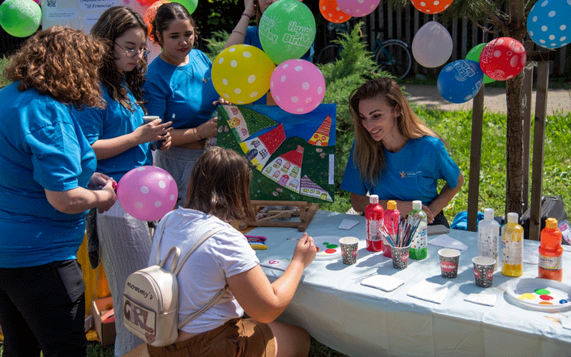 Outdoor arts and crafts booth at a festival: volunteers in blue shirts help a child paint a colorful board as balloons float overhead.