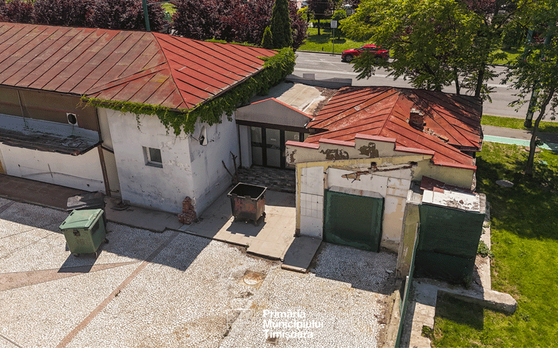 Aerial view of a worn, single-story building with red metal roofs, a gravel yard, and dumpsters outside.