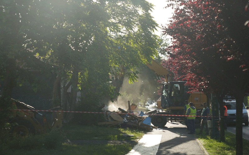 Construction crew and an excavator clearing debris from a street, with red barrier tape and dust in the air.