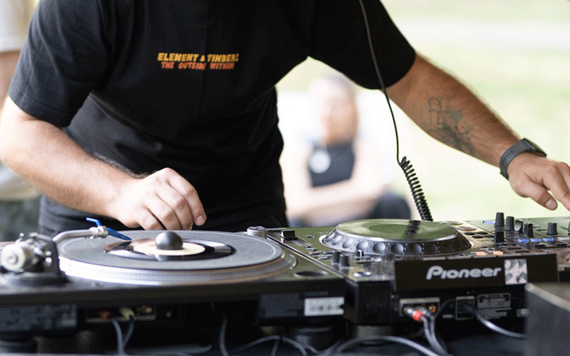 DJ performing outdoors at a vinyl turntable setup with Pioneer mixer and headphones, hands adjusting records and controls on a sunny day.