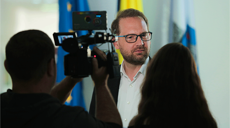 Man with glasses being filmed indoors, camera operator in foreground and flags visible in the background, giving an interview vibe.