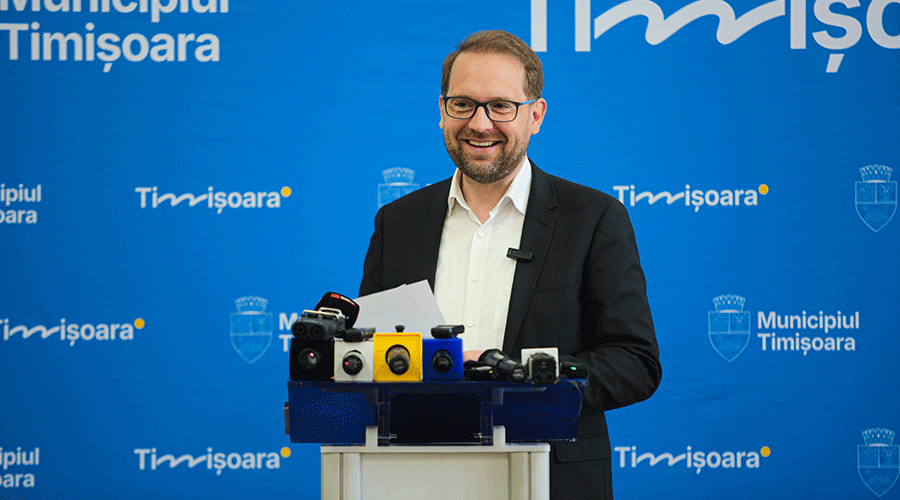 Smiling man in a suit speaks at a podium with multiple microphones during a press conference, blue Timisoara backdrop behind him.