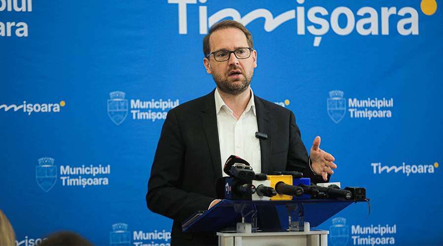 Man in a suit and glasses speaks at a press conference with multiple microphones, set against a blue backdrop with ‘Municipiul Timșoara’ logos.