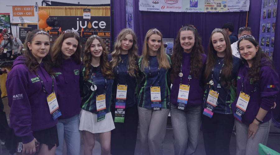 Eight young women in matching purple hoodies with lanyards pose together at a convention booth with banners in the background.