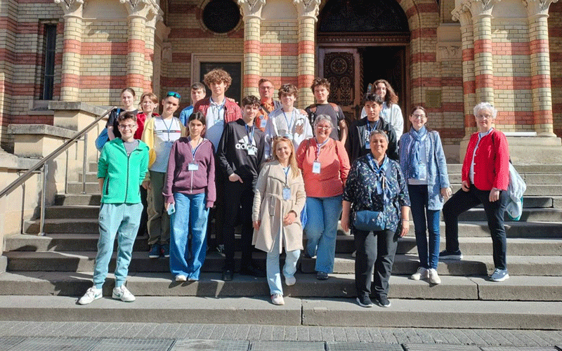 Group of around 18 people posing on the steps of a brick building, wearing lanyards and casual clothing.