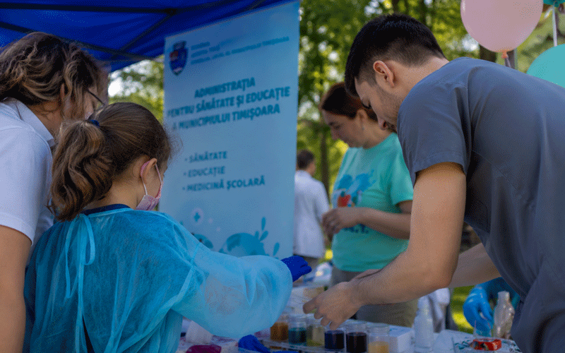 Outdoor education booth at a park: a child in a blue gown and gloves works with a volunteer at a table of colored samples, with a blue banner and balloons in the background.