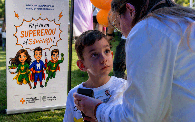 Boy looks up at a woman at an outdoor health event; a poster beside them features cartoon superheroes and Romanian text about health.