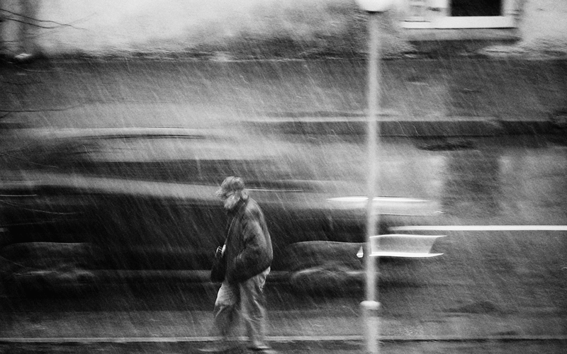 Pedestrian walking in heavy rain along a city street, captured with motion blur.