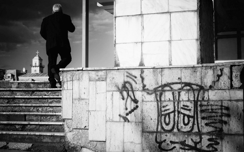 expozitie-carturesti3 - Sursa de Vest Man in a dark suit walking up concrete stairs beside a graffiti-covered wall, in black and white.