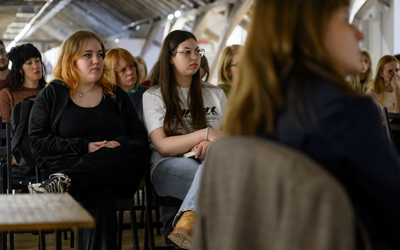 Audience attending a talk: two young women sit in the front row, one with blonde hair in black, the other with glasses, listening attentively in a bright hall.