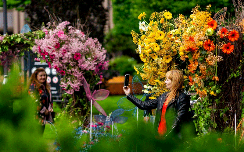 Woman in a black leather jacket takes a selfie with her phone beside a bright flower display in yellows and oranges; another woman watches in the background.