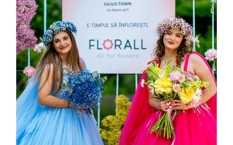 Two women in blue and pink gowns with flower crowns pose holding bouquets at a floral event backdrop reading Floral All for flowers.