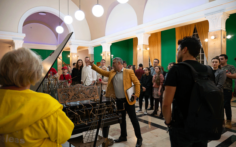 Conductor in a yellow blazer leads a piano performance for a crowd in a grand hall with arches and bright lights.