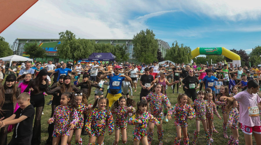 A large group of participants, including children in multicolored outfits, join hands at the start area of an outdoor fun run, with tents and a yellow inflatable arch in the background.