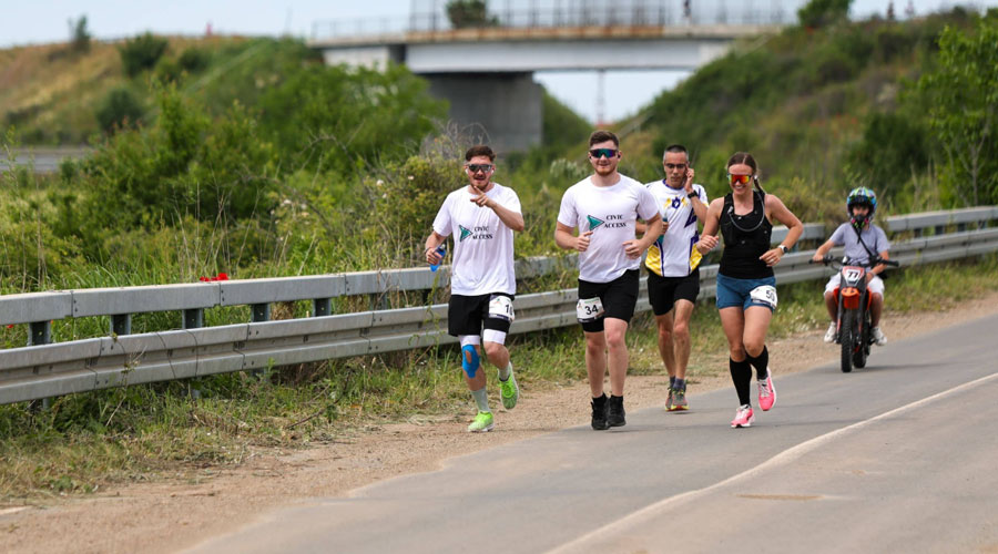 Group of five runners in a road race on a rural highway beside a guardrail, with a child on a small motorcycle behind them in the background.