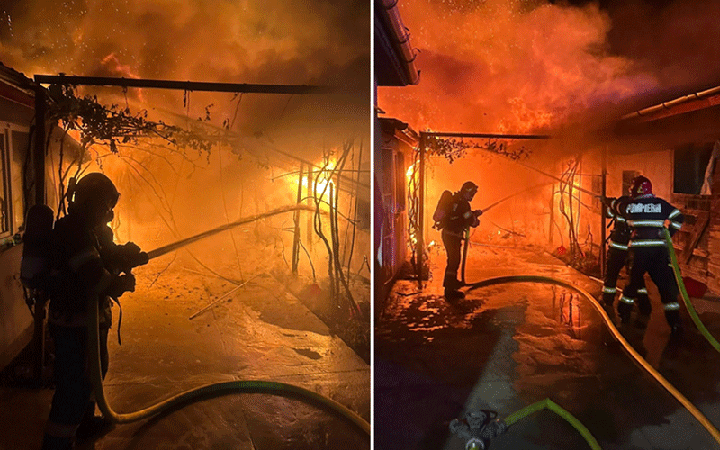 Two firefighters in full turnout gear battle a blaze inside a burning structure, hoses spraying water amid bright orange flames and heavy smoke.