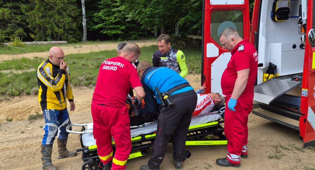 jandarmi montani 2 - Sursa de Vest Paramedics transfer an injured person onto a wheeled stretcher beside a red ambulance outdoors on a dirt path.