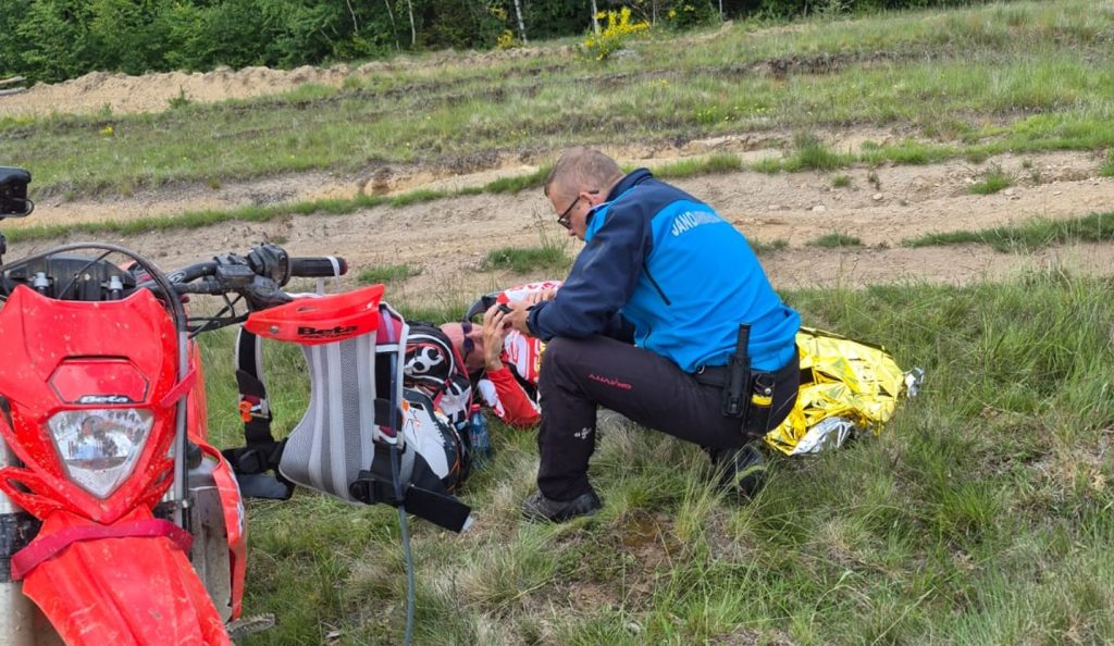 jandarmi montani 3 - Sursa de Vest Rescuer in a blue jacket kneeling beside an injured rider on the grassy roadside, with a red dirt bike nearby and a gold emergency blanket.