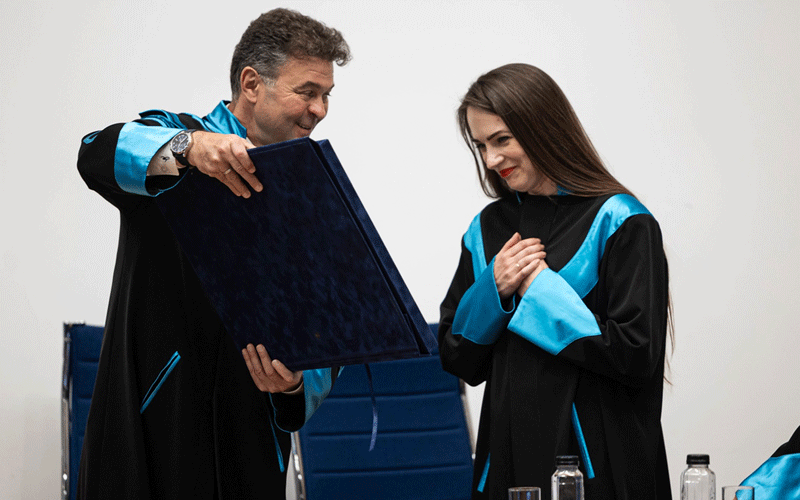 Two graduates in black gowns with blue accents share a moment as one passes a large certificate to the other in a bright room