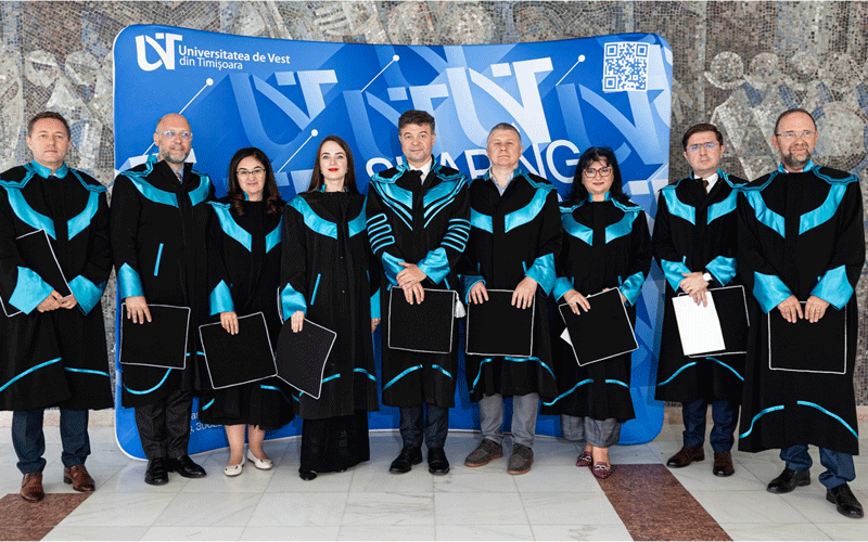 Ten graduates in black robes with teal trim stand in a row, each holding diploma covers, in front of a blue university banner.