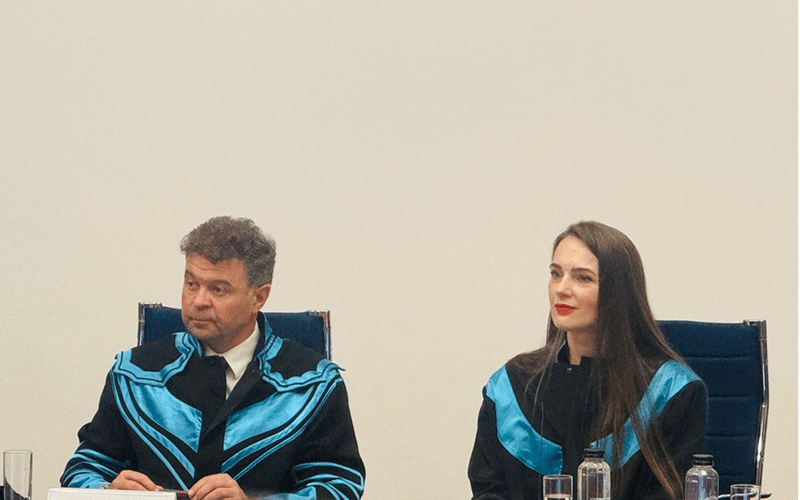 Two people in blue and black academic robes sit at a table with water bottles in a bright room.