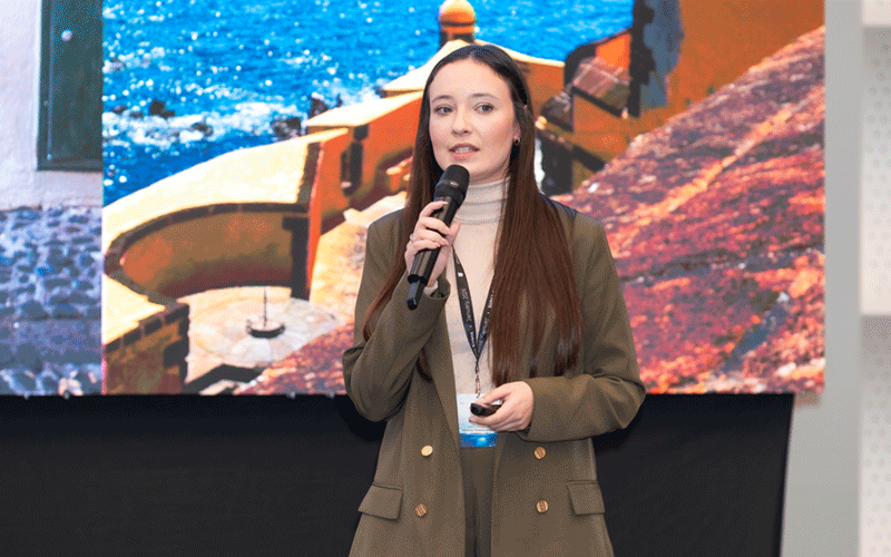 Woman in a green blazer speaks into a handheld microphone on stage, with a large screen showing a coastal scene behind her.