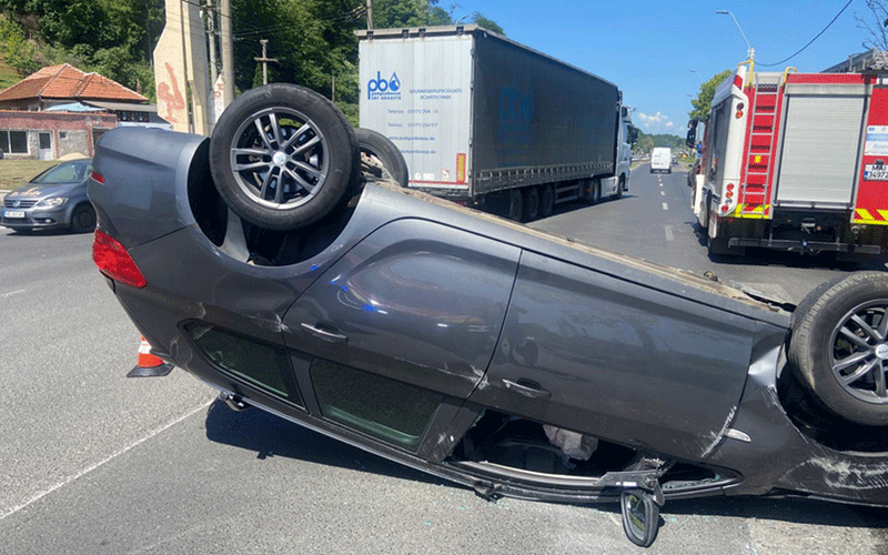 Gray car is overturned on its roof in the middle of a street, wheels in the air, with emergency vehicles and a truck in the background.