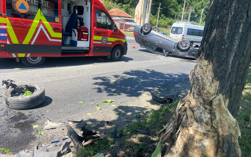 masina-rasturnata2 - Sursa de Vest Red emergency vehicle at the scene of a highway accident; a silver car is flipped on its roof nearby, with debris and a discarded tire on the road.