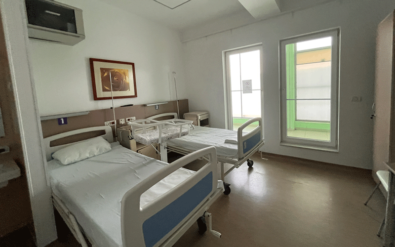 Two hospital beds in a bright patient room with white linens, blue headboard panels, and bedside tables near a large window.
