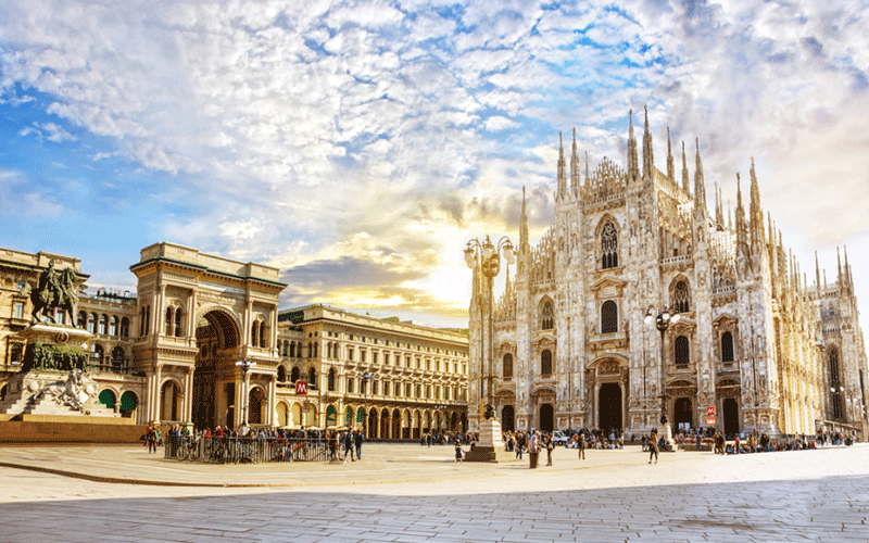 Milano: the ornate Milan Cathedral (Duomo) with the Galleria Vittorio Emanuele II arcaded building to the left, under a partly cloudy sky with people in the square.