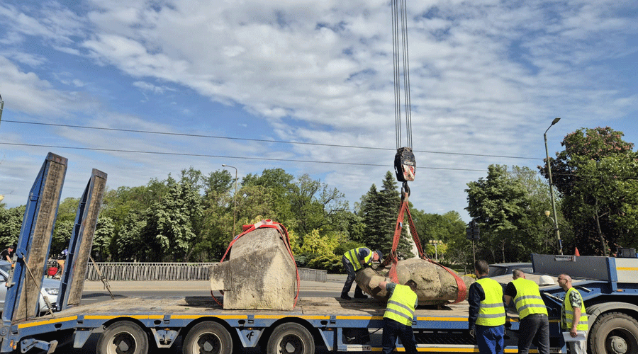 Crane lifts a large boulder onto a blue flatbed trailer as workers in safety vests supervise on a sunny day.