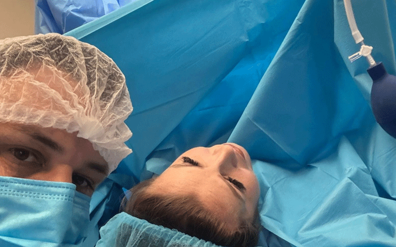 Surgeon wearing a blue surgical cap and mask beside a patient lying on an operating table covered with blue sterile drapes.
