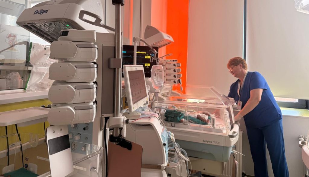neonatologie 3 - Sursa de Vest Nurse in blue scrubs tending a newborn in an incubator with multiple medical monitors in a neonatal unit.