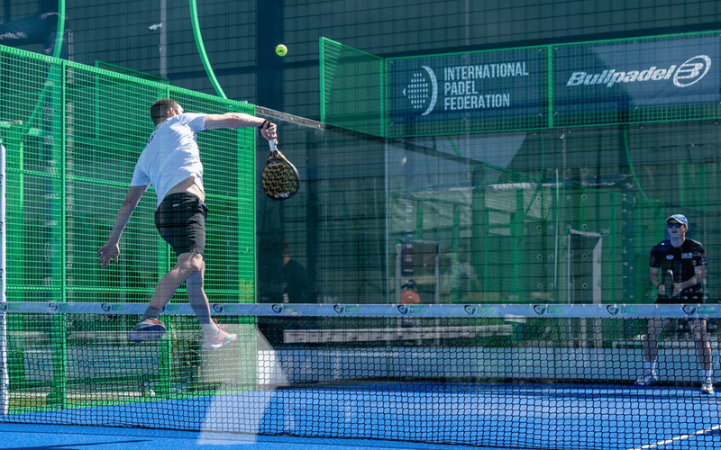 Male padel player in a white shirt and black shorts leaps to strike the ball with a racket on a blue court, green fencing and a net in the foreground, with an observer on the right and banners for the International Padel Federation in the background.