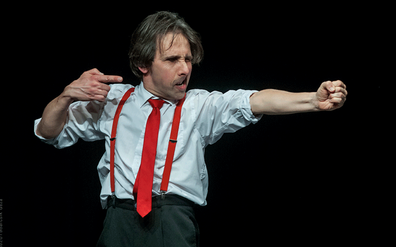 Man in a white shirt, red tie, and red suspenders delivers a dramatic punch on stage against a dark background.