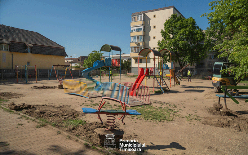 Playground under construction with colorful slides and fences, a worker nearby, and apartment blocks in the background.