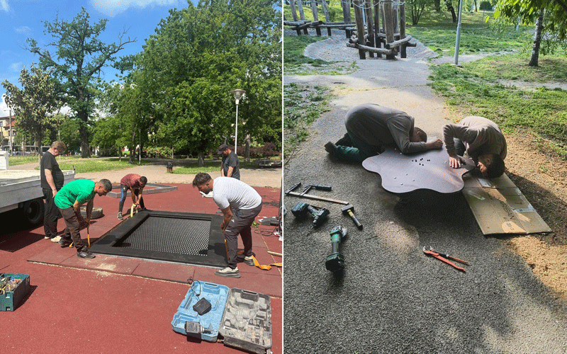 Several workers assemble a modular metal base for a playground on a park court, with toolboxes and equipment nearby.