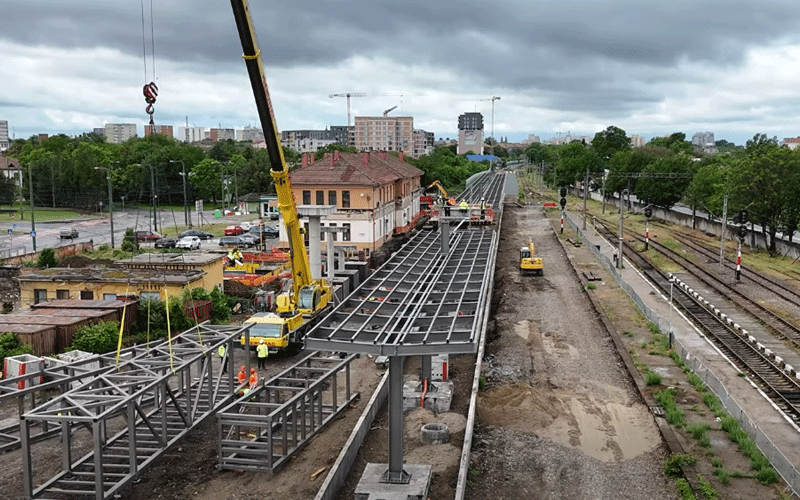 Construction site for a new railway platform with steel framework, a large crane overhead, and workers assembling the structure beside active tracks.