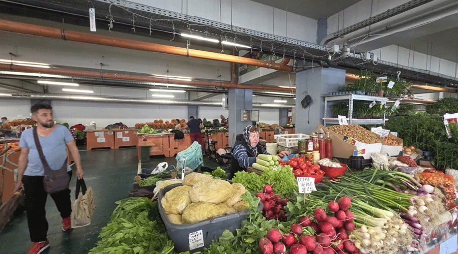 Indoor market scene with a man carrying a bag walking past stalls of fresh produce; a vendor behind a colorful display of vegetables.