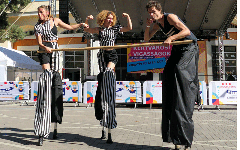 Three stilt performers in black-and-white striped outfits balance on a horizontal bar on an outdoor stage at a festival.