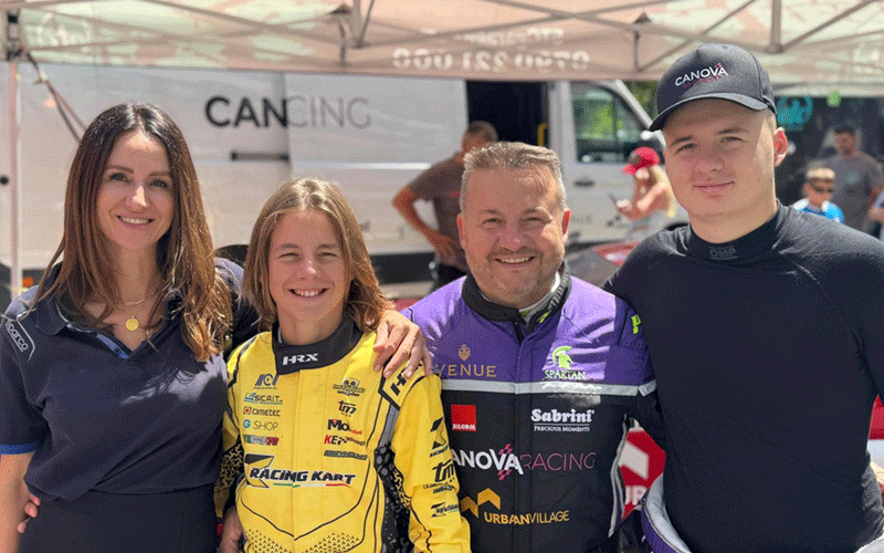 raliu2 - Sursa de Vest Four people posing for a photo at a motorsport event under a canopy; two in racing suits (yellow and purple) stand between a woman in navy and a man in black.
