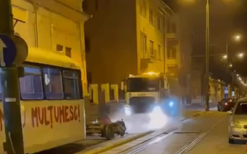 Nighttime urban street scene with a front-facing bus and a person lying on the curb beside tram tracks, headlights and streetlights creating glare.