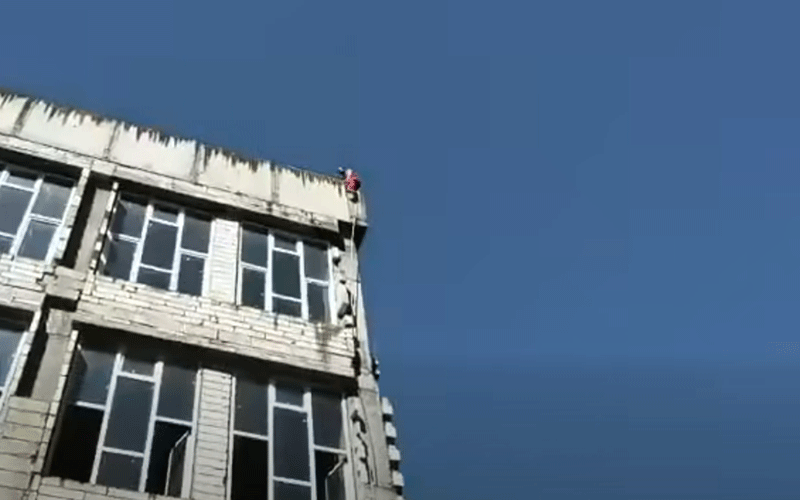 Person wearing red stands on the edge of a rooftop ledge on a partially finished concrete building against a clear blue sky.