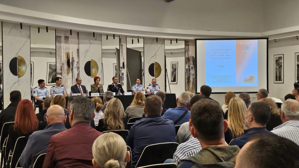 Panel discussion at a formal conference with six speakers seated at a long table, microphones in front, audience facing them, a blue projection slide on the right.