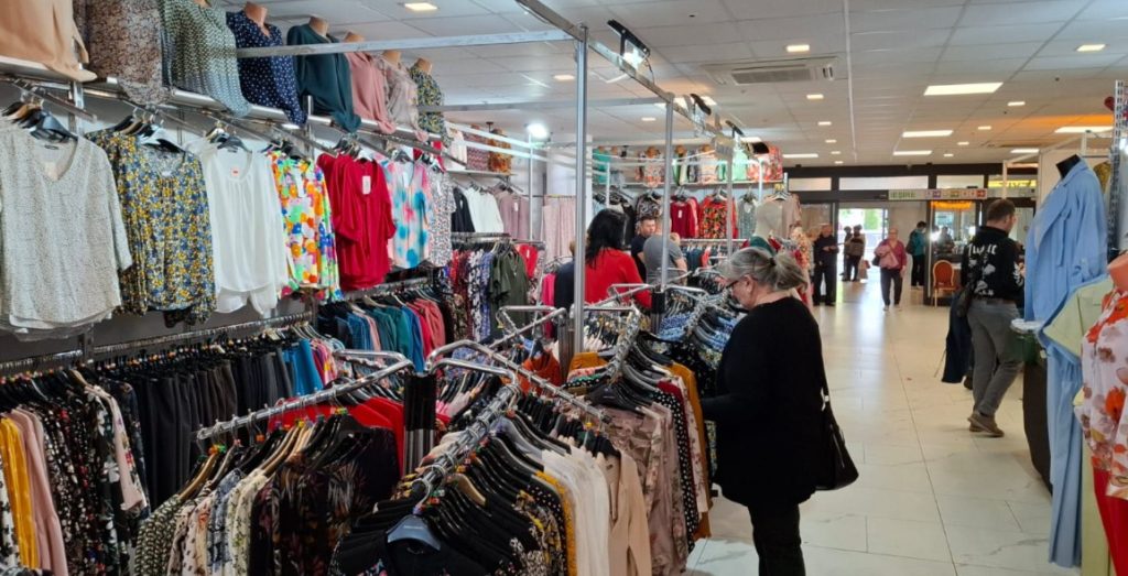 sic 4 - Sursa de Vest Shoppers browse clothing racks in a bright indoor store filled with colorful tops and dresses.
